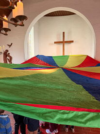 Buntes Schwungtuch, darunter mehrere Kinder in einer Kirche mit Holzkreuz und Kerzenleuchter