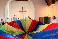 Mehrere Erwachsene stehen in einem Kirchenraum um ein großes, buntes Schwungtuch, im Hintergrund ein Kreuz an der Wand