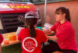 Zwei Personen in roten T-Shirts mit DLRG-Logo sitzen an einem Holztisch unter einem roten Pavillon vor einem DLRG-Fahrzeug