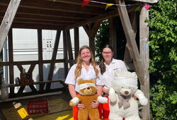 Zwei Frauen in orange-weißer Arbeitskleidung stehen vor einer Holzhütte und halten große Teddybären, eine davon mit Verband am Kopf.