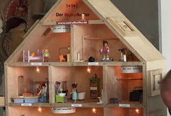 Holz-Puppenhaus mit mehreren beleuchteten Zimmern und kleinen Figuren in verschiedenen Alltagsszenen; ein Zimmer im Erdgeschoss ist verraucht