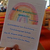 Hand hält ein Programmheft mit einem Regenbogen und dem Text 'Du bist wunderbar gemacht' in einer Kirche mit Holzbänken und einer Holzdecke im Hintergrund