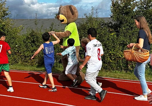 Kinder laufen auf einer roten Laufbahn neben einer Person im Bärenkostüm und einer Frau mit einem Korb