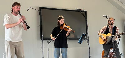 Drei Musiker spielen Klarinette, Geige und Gitarre vor einem großen Bildschirm in einem hellen Raum