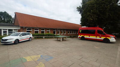 Rotes Einsatzfahrzeug und weißer Kombi vor langgestrecktem Backsteingebäude mit rotem Ziegeldach auf gepflastertem Hof mit bemalten Spielfeldern