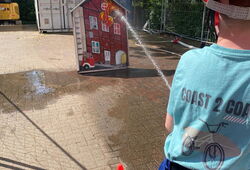 Kind mit oranger Schutzhelmmütze und blauem T-Shirt spritzt mit Wasserschlauch auf ein aufgestelltes Modell eines roten Hauses