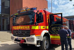 Rotes Feuerwehrfahrzeug mit geöffnetem Seitentür und zwei Personen daneben vor modernem Gebäude