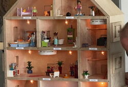 Holz-Puppenhaus mit mehreren beleuchteten Zimmern und kleinen Figuren in verschiedenen Alltagsszenen; ein Zimmer im Erdgeschoss ist verraucht