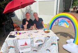 Zwei Personen sitzen an einem mit Regenbogenmuster bedeckten Tisch unter einem roten Sonnenschirm neben einem aufblasbaren Regenbogen mit dem Schild 'Ein Segen für dich'