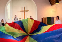 Mehrere Erwachsene stehen in einem Kirchenraum um ein großes, buntes Schwungtuch, im Hintergrund ein Kreuz an der Wand