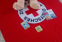 Großer Teddybär mit T-Shirt sitzt auf roter Decke mit weißem Kreis und rotem Kreuz sowie der Aufschrift Deutsches Rotes Kreuz