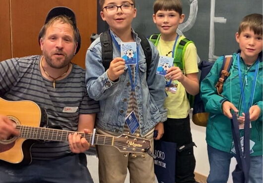 Drei Jungen mit Rucksäcken stehen vor einer Tafel, LiederLukas sitzt daneben und spielt Gitarre