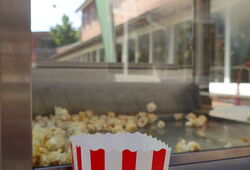Popcorntüte mit roten Streifen und grünem Logo vor einer Popcornmaschine
