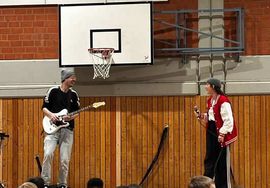 Zwei Personen in Sporthalle vor Basketballkorb, eine spielt E-Gitarre, die andere hält Mikrofon und trägt rote Collegejacke
