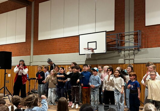 Kinder stehen in einer Turnhalle vor einem Basketballkorb auf einer Bühne und singen oder sprechen gemeinsam, links steht eine Person mit Mikrofon