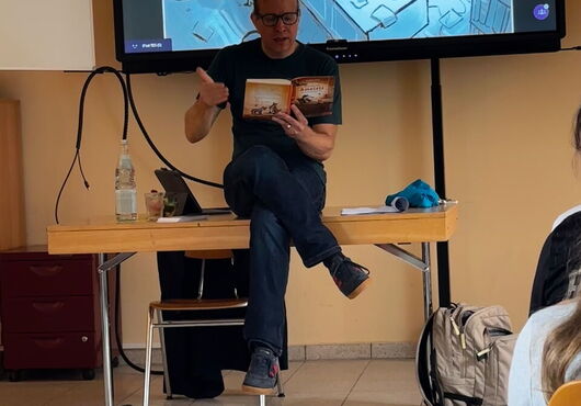 Matthias Kröner sitzt auf Tischkante vor Bildschirm mit geöffneter Buchseite und liest vor, im Hintergrund schwarz-weiße Linienzeichnung eines Zimmers mit Schreibtisch und Stuhl