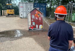 Rückansicht einer Person mit orangefarbenem Schutzhelm vor einem kleinen Modellhaus auf einem gepflasterten Platz mit Containern und Bagger im Hintergrund
