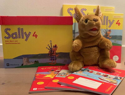 Plüschkänguru sitzt vor mehreren gelben und roten Lernheften und einer gelben Box mit der Aufschrift 'Sally 4'