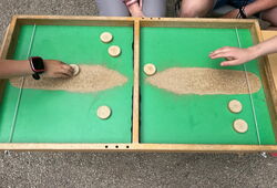 Zwei Kinder spielen an einem Holztisch mit grünem Spielfeld und Holzscheiben, in der Mitte liegt eine Sandspur