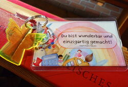 Schlüsselanhänger mit gelbem Engel liegt auf einer roten Bank, daneben eine Karte mit der Aufschrift 'Du bist wunderbar und einzigartig gemacht!'