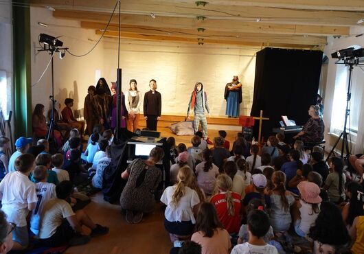 Kinder sitzen auf dem Boden und schauen eine Theateraufführung mit mehreren Schauspielern auf einer Bühne in einem Raum mit Beleuchtung und schwarzem Vorhang