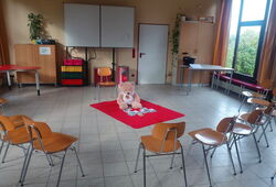 Leerer Klassenraum mit Stühlen im Halbkreis um einen roten Teppich mit einem großen Teddybär in der Mitte