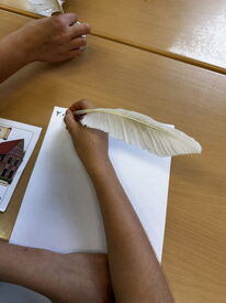 Kind hält eine weiße Feder und schreibt auf ein weißes Blatt Papier auf einem Holztisch