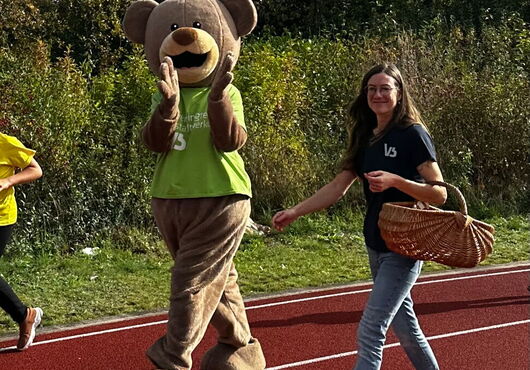 Bär-Maskottchen mit grünem T-Shirt und Person mit Korb laufen über rote Laufbahn
