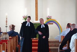 Menschen sitzen in einer Kirche und hören zwei Pastorinnen am Altar zu