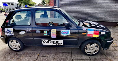 Kleiner schwarzer Pkw mit mehreren bunten Aufklebern an der Seite und auf der Motorhaube, darunter ein Aufkleber mit der Aufschrift 'KulTour mobil'