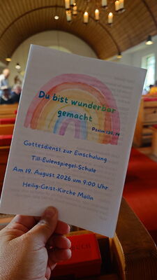 Hand hält ein Programmheft mit einem Regenbogen und dem Text 'Du bist wunderbar gemacht' in einer Kirche mit Holzbänken und einer Holzdecke im Hintergrund