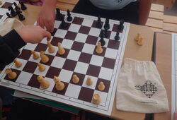 Zwei Personen spielen Schach an einem Schachbrett mit weißen und schwarzen Figuren auf einem Holztisch, daneben ein Beutel mit Schachlogo