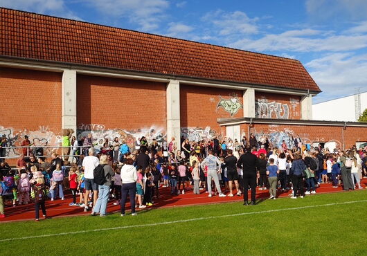 Große Gruppe von Kindern und Erwachsenen steht auf rotem Sportplatz vor einer mit Graffiti bedeckten Backsteinmauer unter blauem Himmel
