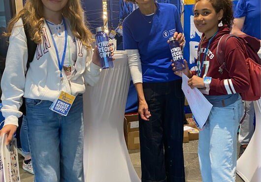 Zwei Mädchen und eine Frau stehen an einem Stehtisch, die Mädchen tragen Jeans und Sweatshirts, die Frau ein blaues T-Shirt mit Aufdruck