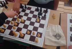 Zwei Personen spielen Schach an einem Schachbrett mit weißen und schwarzen Figuren auf einem Holztisch, daneben ein Beutel mit Schachlogo