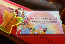 Schlüsselanhänger mit gelbem Engel liegt auf einer roten Bank, daneben eine Karte mit der Aufschrift 'Du bist wunderbar und einzigartig gemacht!'