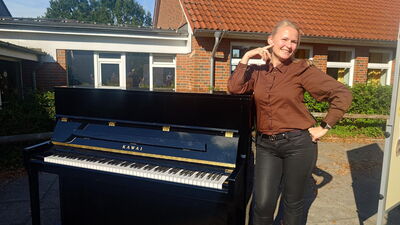Eine Frau steht lächelnd neben einem schwarzen Klavier auf dem Schulhof. Hinter ihr sind das rote Backsteingebäude der Schule und Bäume zu sehen. Das Foto gehört zur musikalischen Begrüßung durch den Schulchor.