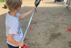 Junge mit weißem T-Shirt und dunkler Hose hält Lacrosse-Schläger auf einem Spielfeld mit roten Markierungshütchen
